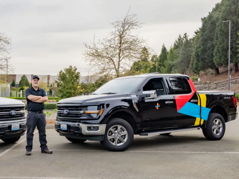 Two branded pickup trucks, one white and one black, parked side by side with a uniformed worker standing between them.