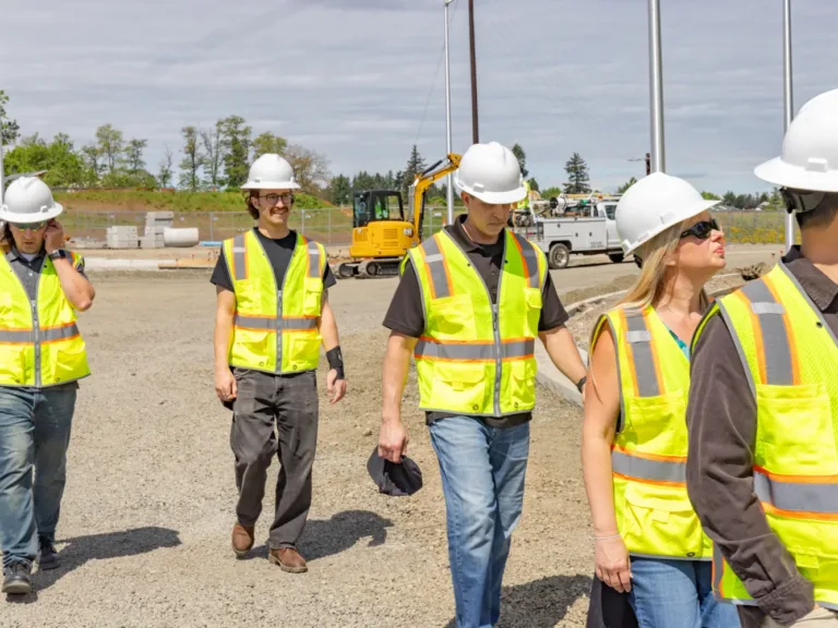 MacKay field crew in high-visibility vests and hard hats walking a construction site, with a yellow excavator, utility truck, and graded gravel in the background.