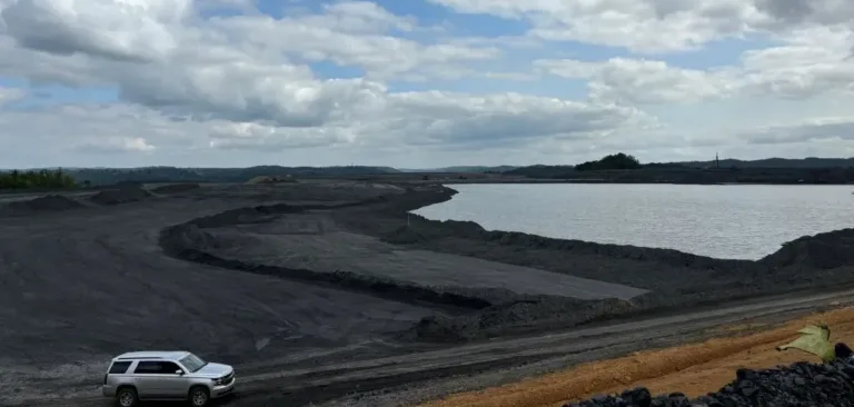 Open-pit mining site with a large water-filled excavation, dark earthworks, and an SUV in the foreground beneath a cloudy sky