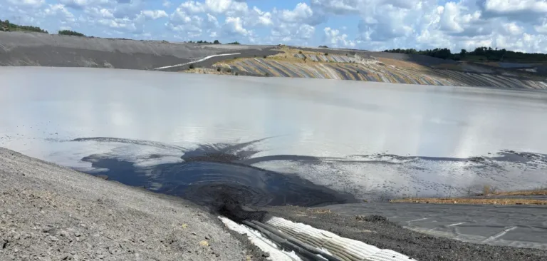Lined industrial settling pond with gray water, dark sediment, and a large corrugated drainage pipe in the foreground