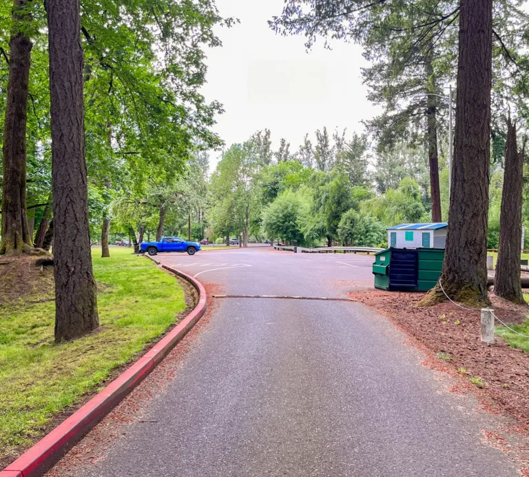 Paved park road winding through tall trees, with a blue pickup truck, parking area, and green dumpster
