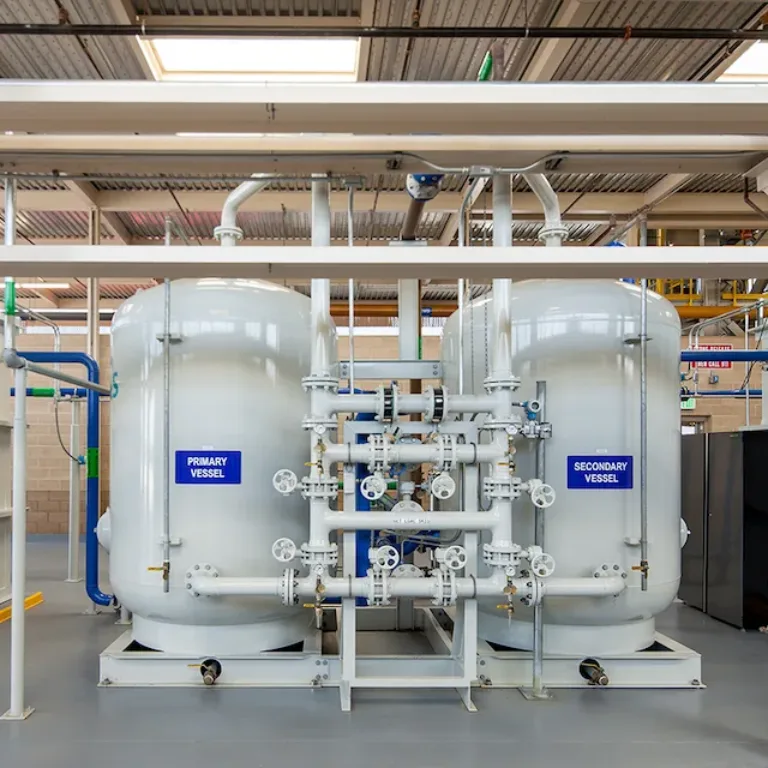 Industrial water-treatment system with two large tanks labeled “PRIMARY VESSEL” and “SECONDARY VESSEL,” connected by pipes and valves