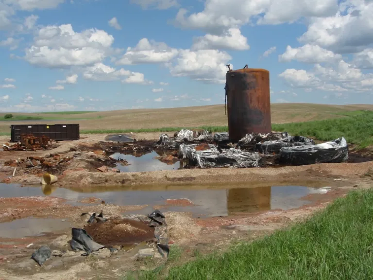 Rusted industrial tank and tarp-covered debris beside oily puddles in a rural field