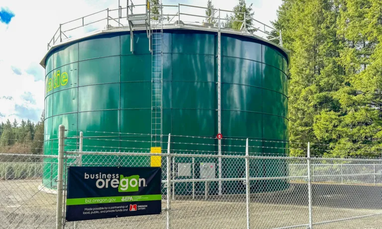 Large green cylindrical water tank behind a security fence, bearing a partial “...alie” label and a Business Oregon sign