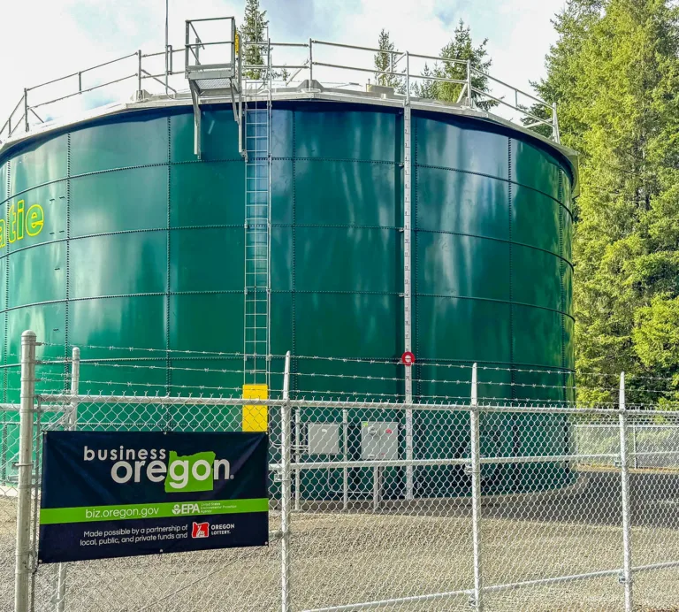 Large green cylindrical water tank behind a security fence, bearing a partial “...alie” label and a Business Oregon sign