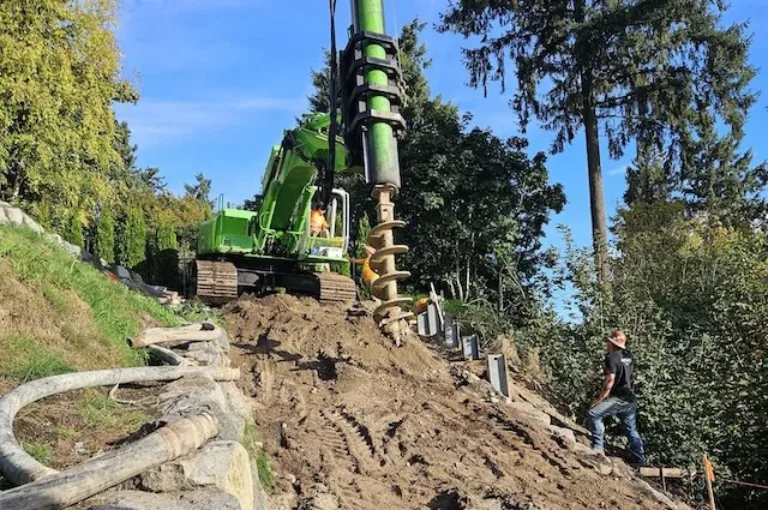 Excavator with a large auger drills into a dirt hillside while a worker stands nearby