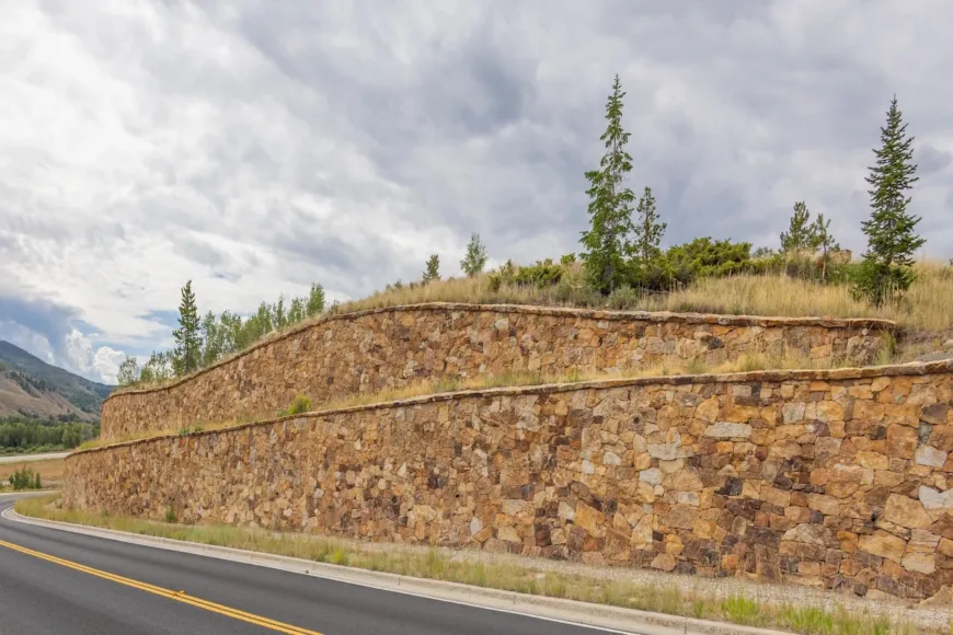 Tiered stone retaining walls beside a two-lane road, with grassy slopes, evergreen trees, and mountains under a cloudy sky
