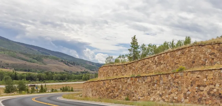 Curving mountain road beside terraced stone retaining walls, with wooded hills and a cloudy sky in the background