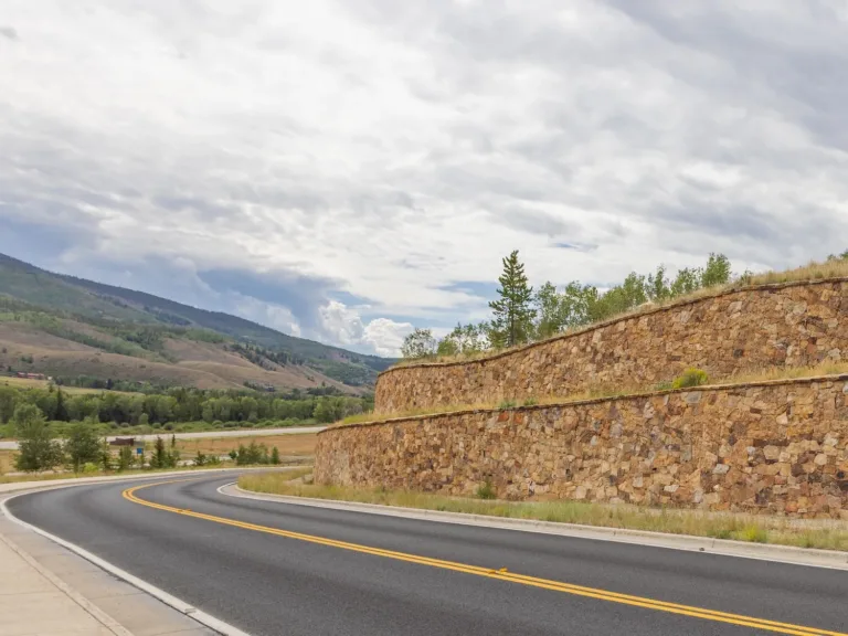 Tiered stone retaining walls beside a two-lane road, with grassy slopes, evergreen trees, and mountains under a cloudy sky