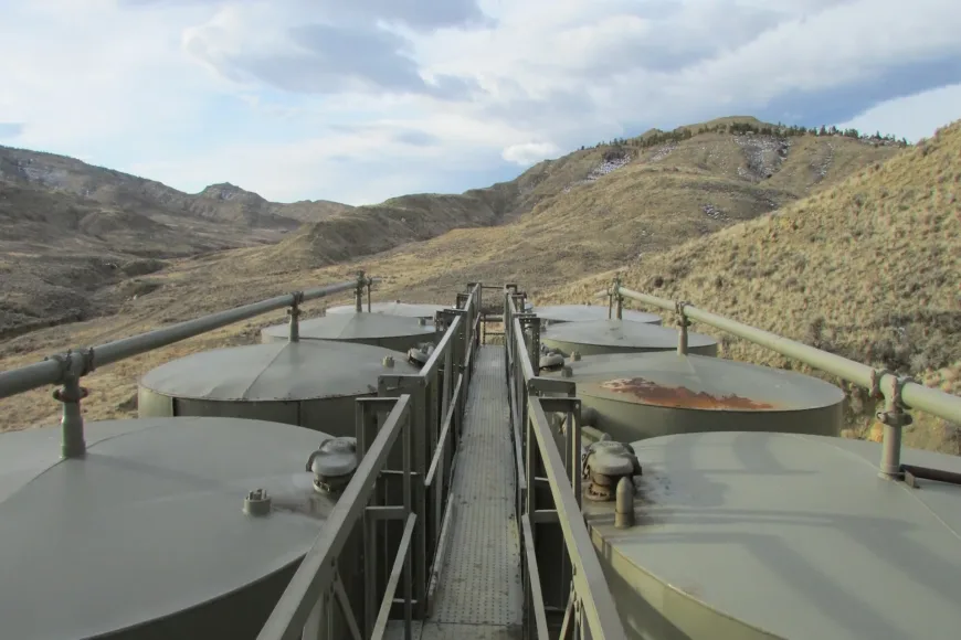 Large cylindrical storage tanks connected by pipes and a central catwalk in a barren, rolling mountain landscape