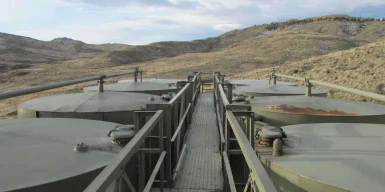 A narrow metal walkway crosses large industrial storage tanks in a barren, hilly landscape