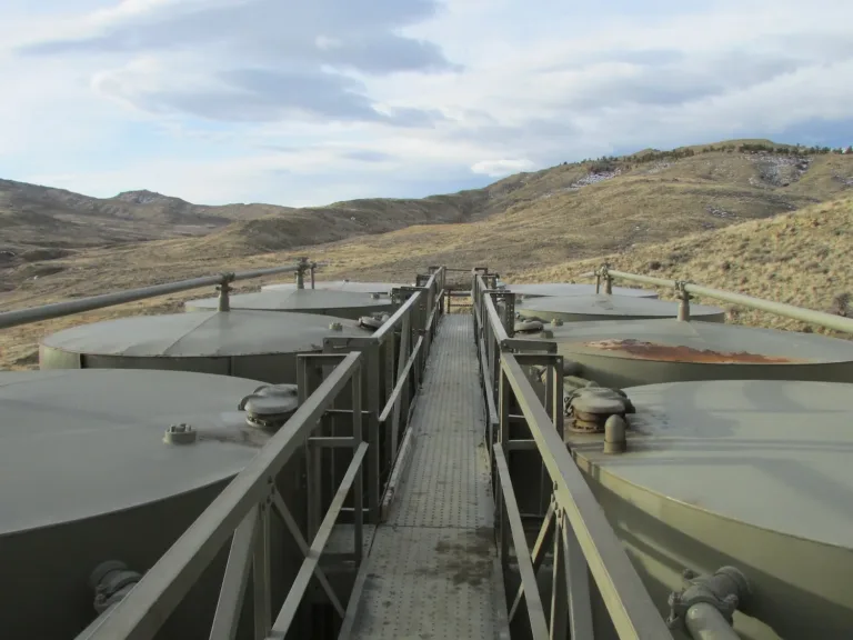 Large cylindrical storage tanks connected by pipes and a central catwalk in a barren, rolling mountain landscape