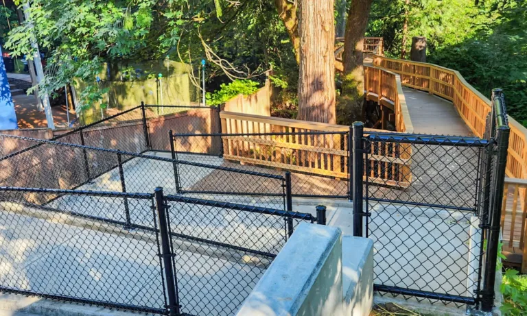 Accessible wooden boardwalk winding through a wooded area beside a fenced concrete walkway