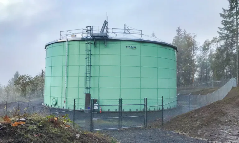 Large green cylindrical storage tank with rooftop access equipment, enclosed by a chain-link fence in a wooded area