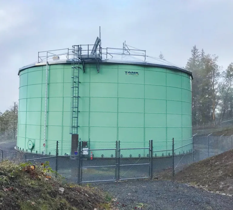 Large green cylindrical storage tank with rooftop access equipment, enclosed by a chain-link fence in a wooded area