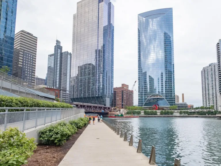 Chicago Riverwalk with pedestrians beside the river, surrounded by glass-and-steel skyscrapers and a nearby bridge