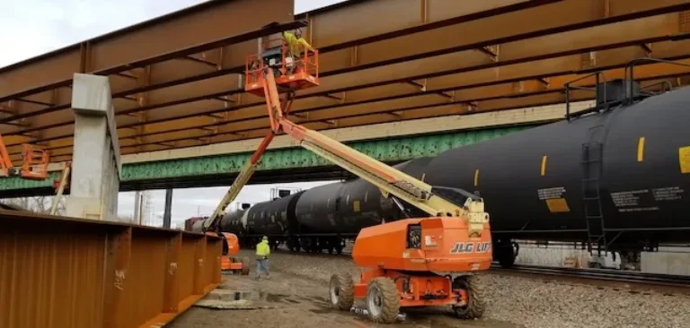 Workers use an orange boom lift beneath a bridge while black tanker railcars pass alongside