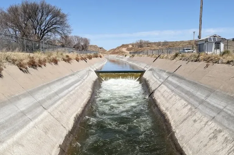 Concrete-lined canal with water cascading over a small weir, bordered by dry grass and leafless trees