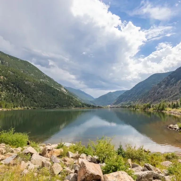 Mountain valley surrounding a calm lake, with forested slopes, rocky shoreline, and dramatic clouds reflected in the water