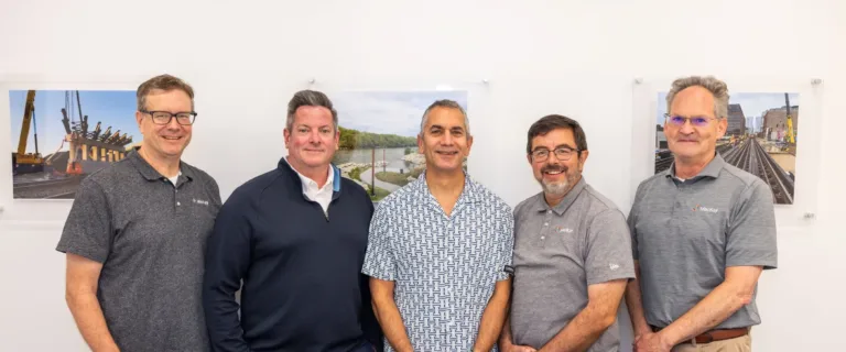 Five men posing together in an office, with construction and infrastructure photos displayed behind them