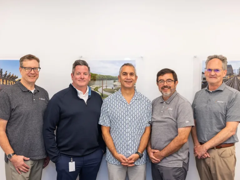 Five men posing together in an office, with construction and infrastructure photos displayed behind them