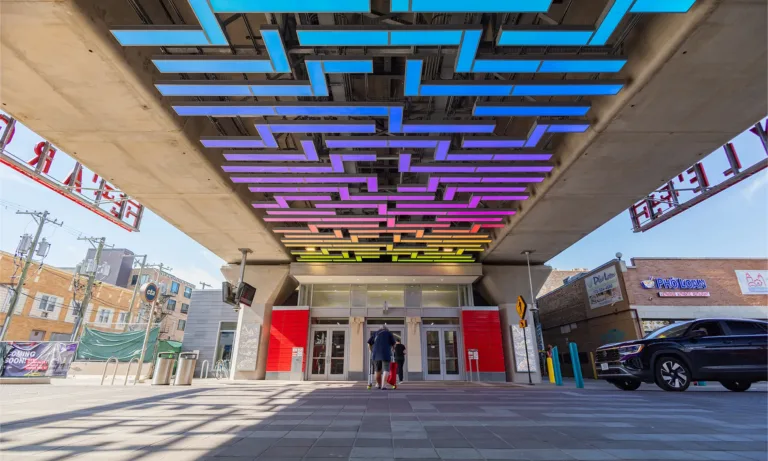 Transit station entrance beneath elevated tracks, with colorful geometric ceiling lights and people entering