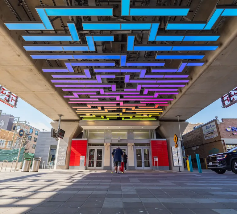 Transit station entrance beneath elevated tracks, with colorful geometric ceiling lights and people entering