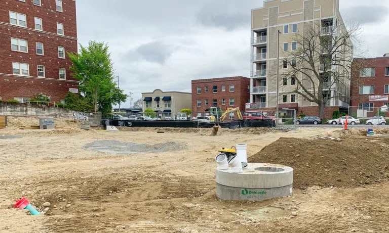 Urban construction site with exposed soil, utility infrastructure, and excavation equipment between surrounding apartment buildings