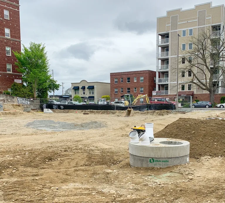 Urban construction site with exposed soil, utility infrastructure, and excavation equipment between surrounding apartment buildings