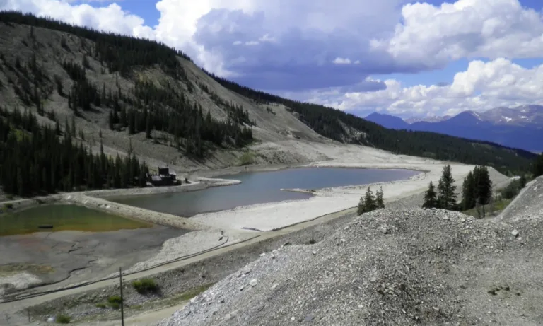 Mountain reservoirs surrounded by evergreen forests and rocky slopes, with snow-capped peaks beneath a cloudy blue sky
