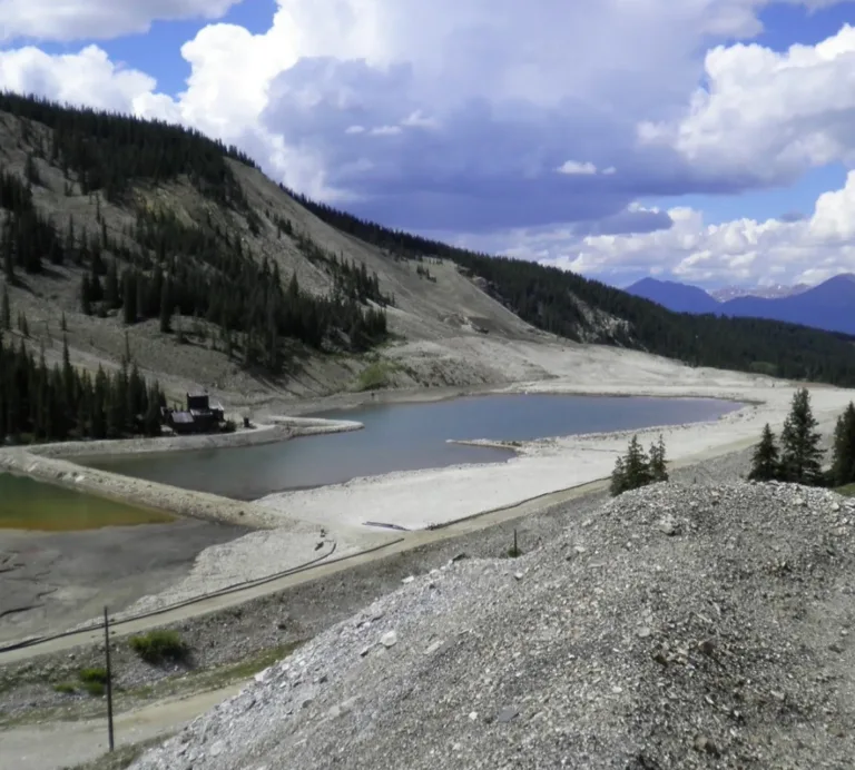 Mountain reservoirs surrounded by evergreen forests and rocky slopes, with snow-capped peaks beneath a cloudy blue sky