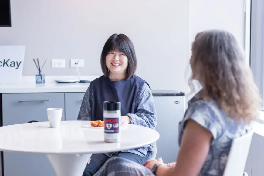 Two people sit across from each other at a table, with one smiling and a drink and snacks between them