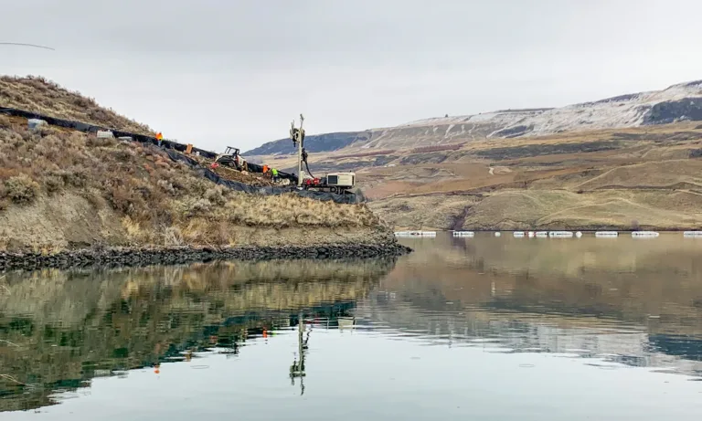 Workers operate drilling equipment on a vegetated slope beside a reservoir, with rolling hills in the background