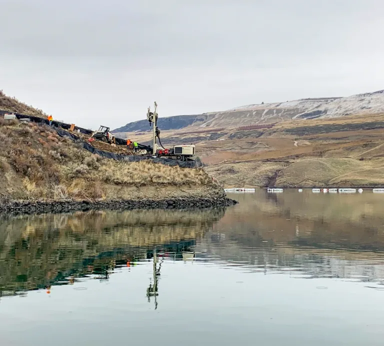 Workers operate drilling equipment on a vegetated slope beside a reservoir, with rolling hills in the background