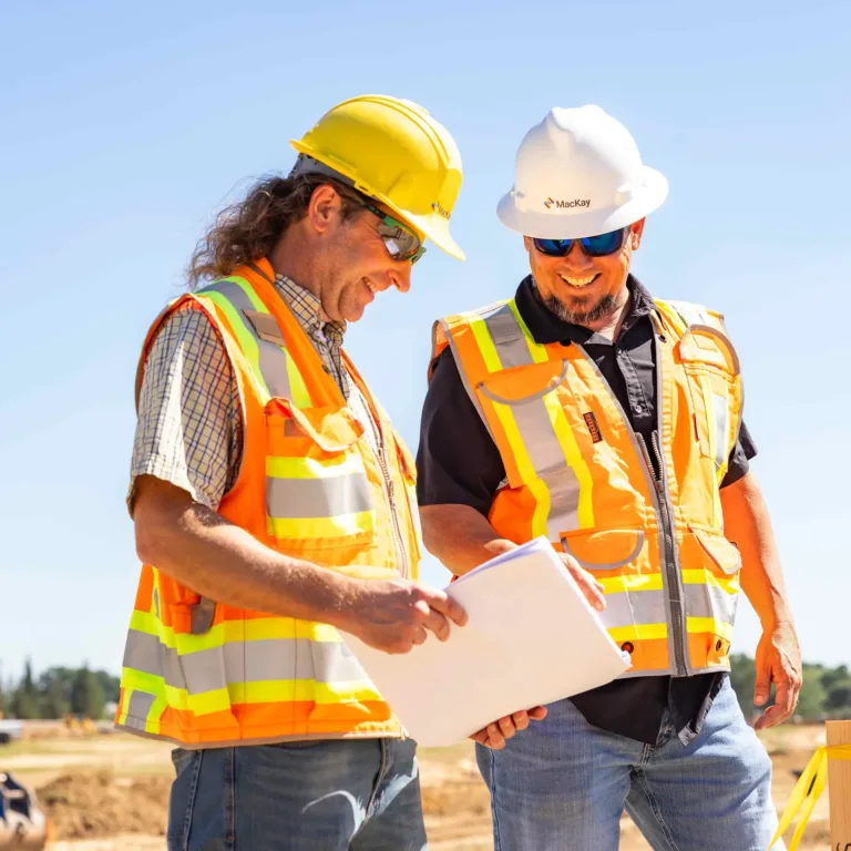 Two construction workers in high-visibility vests and hard hats review blueprints at a construction site