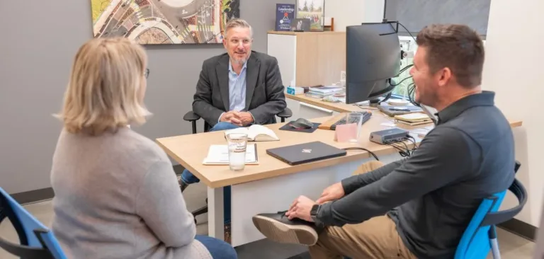 Three people meet in an office around a desk, with one person seated behind a computer monitor