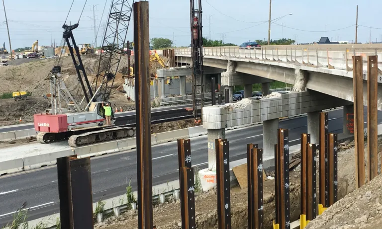 Highway bridge construction with cranes, excavators, concrete supports, and steel sheet piles beside active traffic lanes