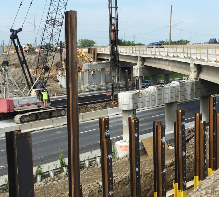 Highway bridge construction with cranes, excavators, concrete supports, and steel sheet piles beside active traffic lanes