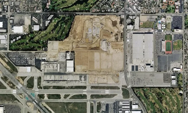 Aerial view of a large construction site surrounded by roads, industrial buildings, a golf course, and sports fields