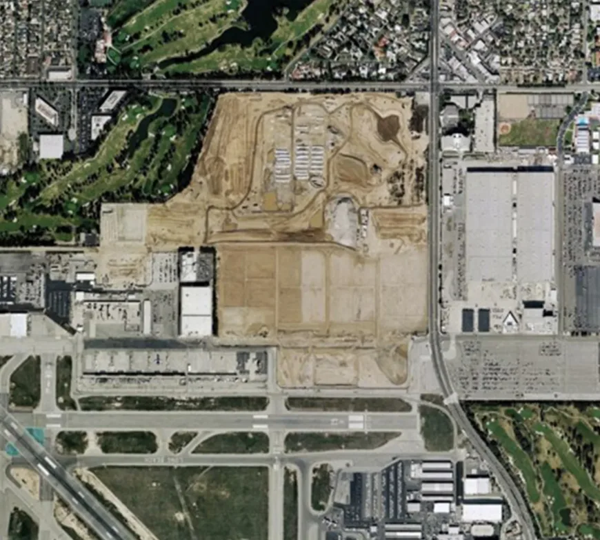 Aerial view of a large construction site surrounded by roads, industrial buildings, a golf course, and sports fields