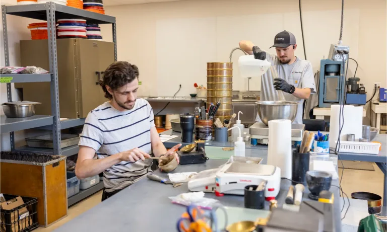 Two workers prepare materials at a manufacturing workbench, with one weighing ingredients and the other pouring from a container