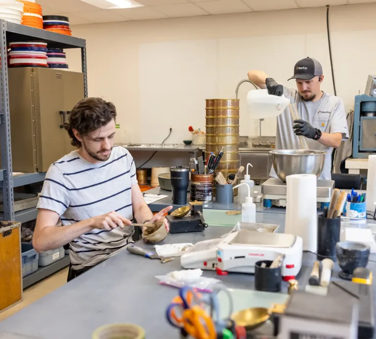 Two workers prepare materials at a manufacturing workbench, with one weighing ingredients and the other pouring from a container