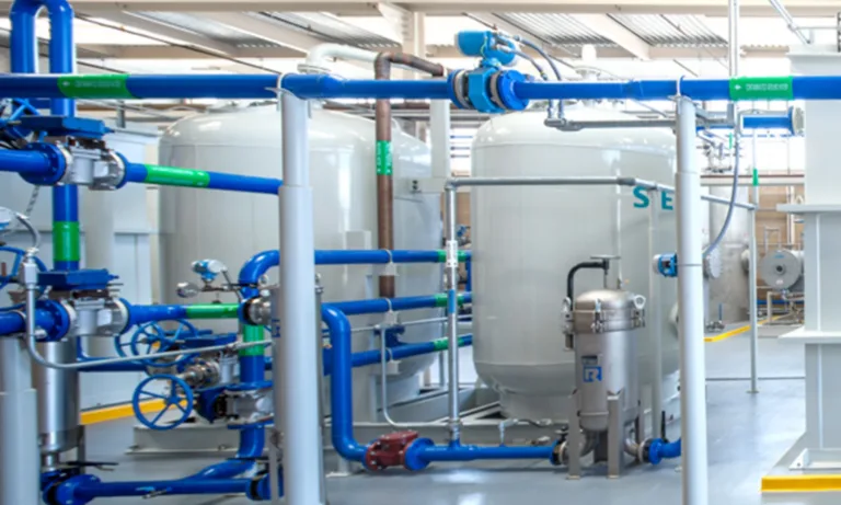 Industrial water-treatment system with blue and green pipes, valves, pumps, and large white storage tanks