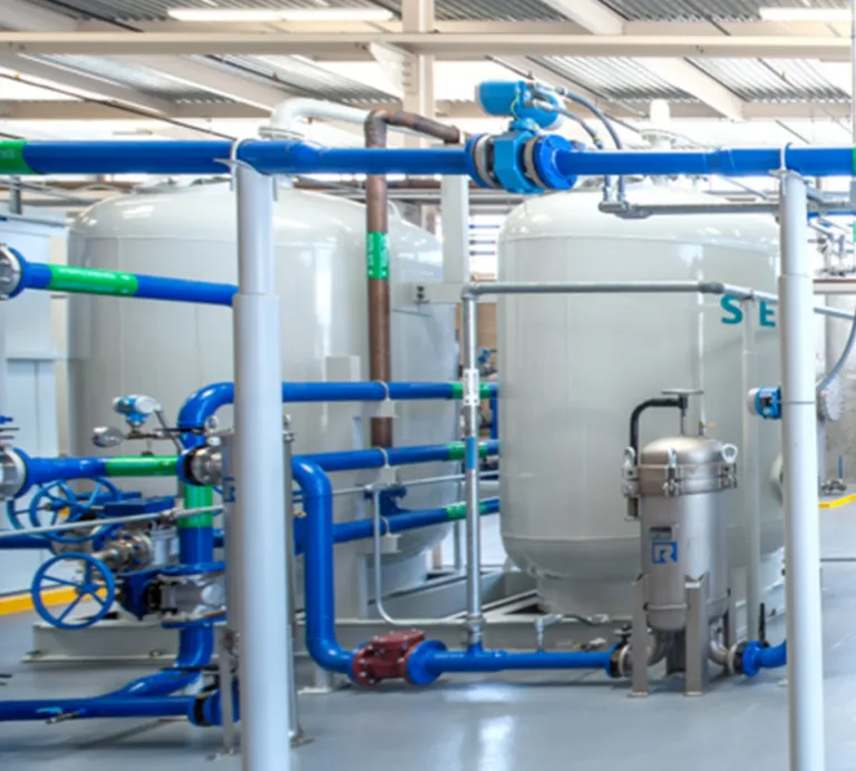 Industrial water-treatment system with blue and green pipes, valves, pumps, and large white storage tanks