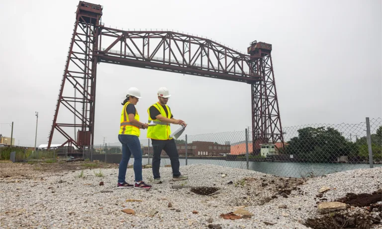 Two engineers in hard hats and safety vests review plans at an industrial site beneath a steel bridge structure
