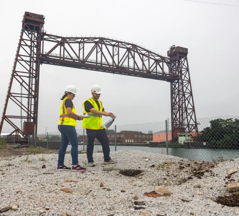 Two engineers in hard hats and safety vests review plans at an industrial site beneath a steel bridge structure