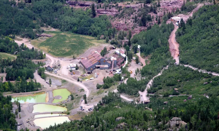 Aerial view of a mining complex with weathered buildings, dirt roads, settling ponds, and forested rocky hillsides