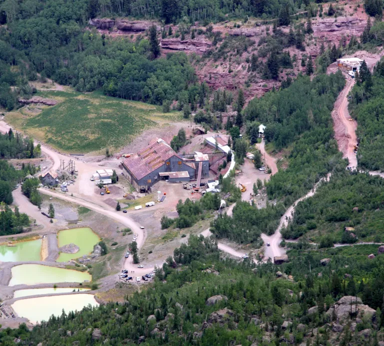 Aerial view of a mining complex with weathered buildings, dirt roads, settling ponds, and forested rocky hillsides