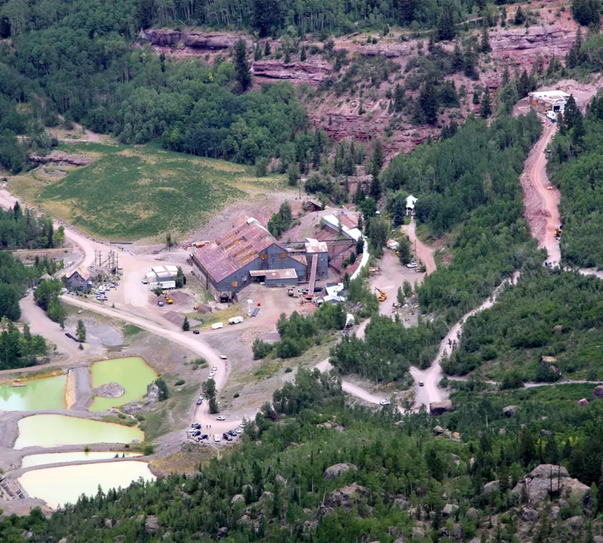 Aerial view of a mining complex with weathered buildings, dirt roads, settling ponds, and forested rocky hillsides