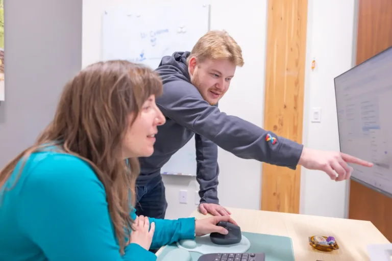 Two colleagues collaborate at a workstation, with one pointing at information on a monitor while the other uses a mouse
